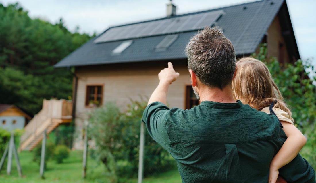 Vater mit Kind auf dem Arm zeigt auf Haus mit Photovoltaik Dach