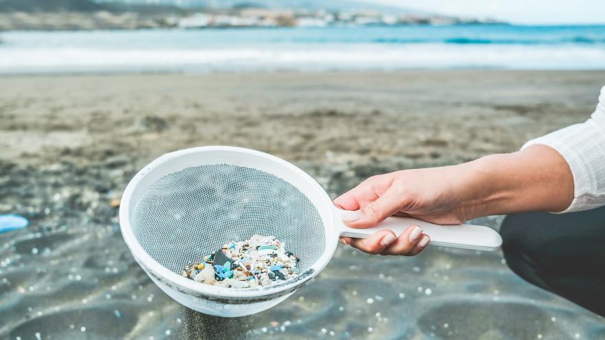 Frau am Strand sammelt mit einem Sieb kleine Plastikteilchen aus dem Sand