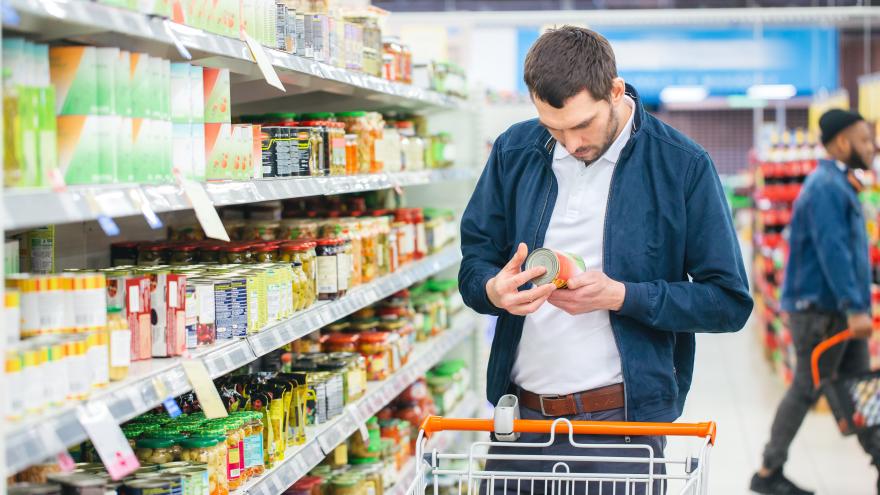 Mann steht mit Einkaufswagen vor einem Supermarktregal und betrachtet die Inhalststoffe eines Produkts