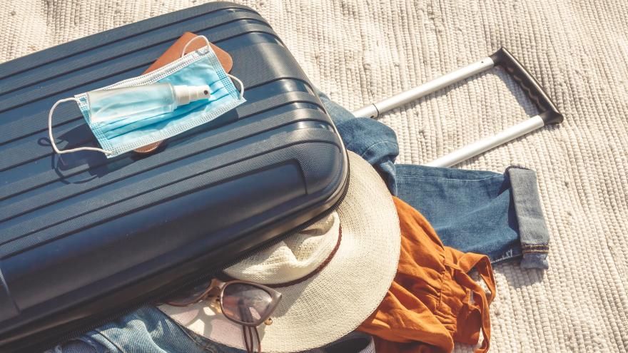 Medizinische Gesichtsmaske und Desinfektionsmittel liegen auf einem blauen Reisekoffer und verschiedenen Kleidungsstücken