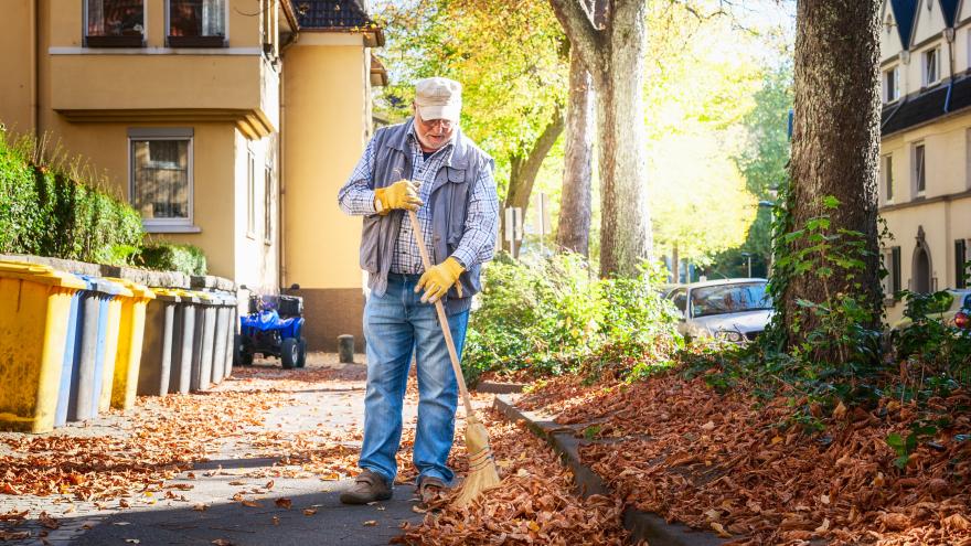 Älter Mann kehrt das Herbstlaub auf dem Gehweg zusammen
