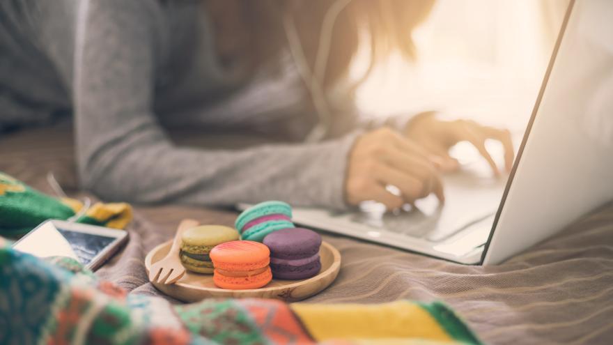 Frau liegt mit Laptop und einem Teller mit Macarons auf dem Bett