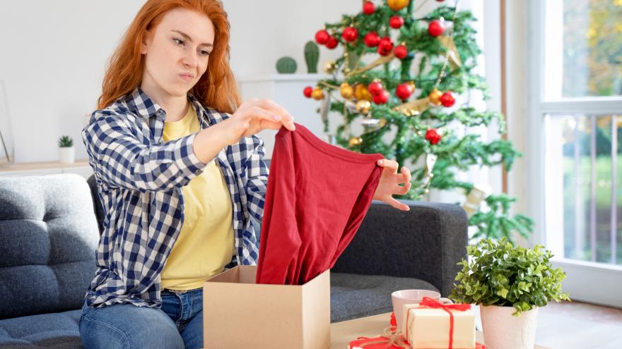 Frau sitzt vor einem Weihnachtsbaum auf der Couch und öffnet ein Geschenk, das ihr nicht gefällt