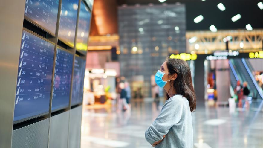Frau mit Gesichtsmaske steht am Flughafen vor einer Anzeigetafel