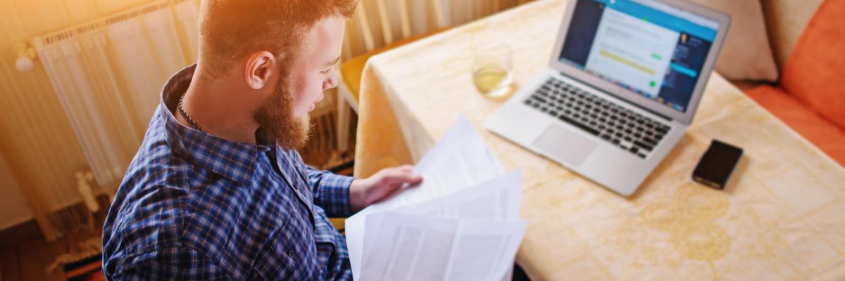 Ein Mann sitzt in seiner Wohnung am Laptop und studiert ausgedruckte Unterlagen