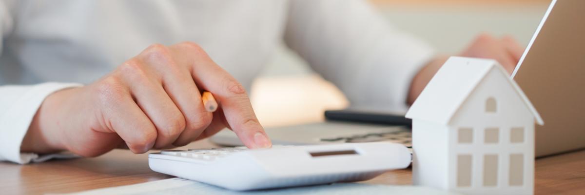 Jemand sitzt an seinem Laptop und rechnet mit einem Taschenrechner. Daneben steht ein kleines Modell eines Hauses.