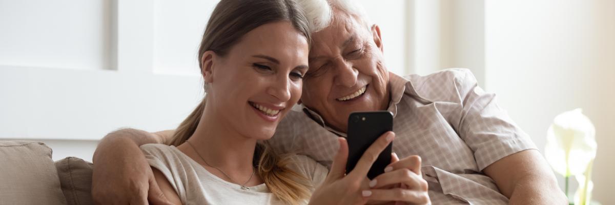 Eine Tochter sitzt mit ihrem Vater auf dem Sofa und sie blicken gemeinsam auf ein Smartphone.