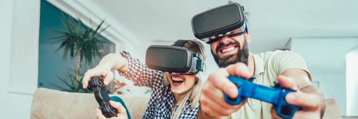 Ein junges Paar sitzt lachend auf einem beigen Sofa und trägt Virtual-Reality-Headsets. Beide halten Gaming-Controller in den Händen und scheinen mitten in einem Videospiel zu sein. Die Frau trägt ein kariertes Hemd und gepunktete Socken, während der Mann ein helles Poloshirt trägt. Vor ihnen auf dem Sofa liegen Kopfhörer und eine Schüssel Popcorn.