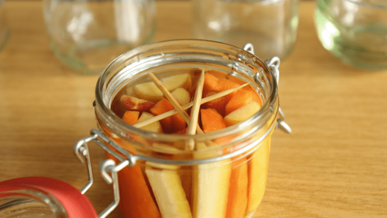 Ein offenes Einmachglas mit Gemüsesticks und Wasser. Holzstäbchen, die zwischen die Glaswände gepresst sind, halten das Gemüse unter der Wasseroberfläche.