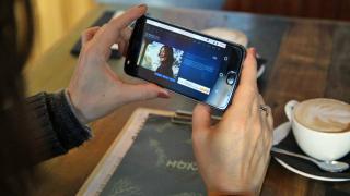 Eine Frau hält in einem Café ein Smartphone in der Hand, auf dem ein Video-Streaming-Dienst zu sehen ist.