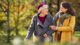 Ältere Frau mit weißem Haar und rotem Barett geht mit junger Frau im herbstlichen Park. Die jüngere Frau stützt sie. Bäume mit bunten Blättern im Hintergrund.