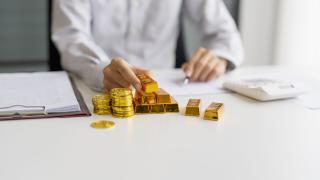 Auf einem Schreibtisch liegen Goldbarren und Münzen, jemand sitz am Schreibtisch mit Taschenrechner