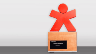 Bundespreis der Stiftung Verbraucherschutz: Ein rote Figürchen, das die Arme wegstreckt und auf einem Holzsockel befestigt ist