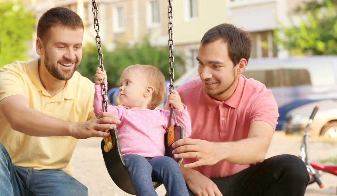 Zwei Männer spielen mit ihrer Tochter, die auf einer Schaukel sitzt.