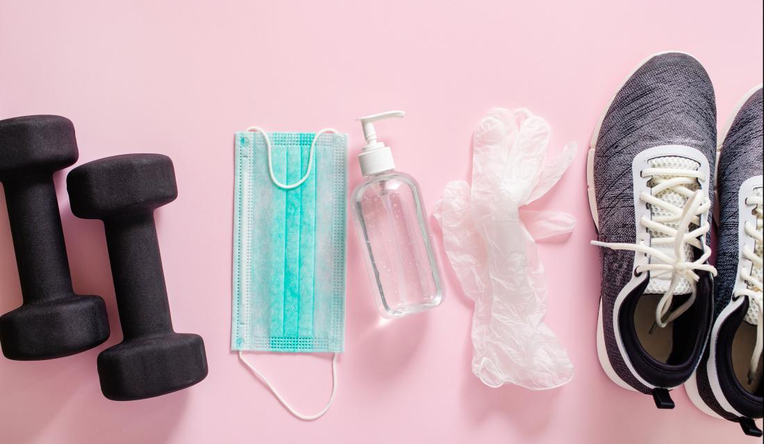 Hantel, Gesichtsmaske, Desinfektionsmittel und Turnschuhe vor rosa Hintergrund