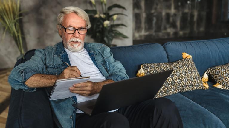 Älterer Mann sitzt mit Laptop auf der COuch und nimmt an einem Webinar teil