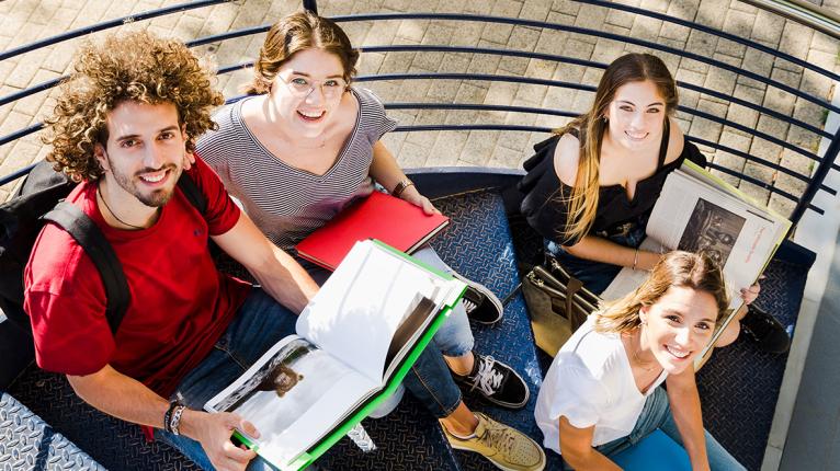 Vier junge Erwachsene sitzen mit Büchern auf einer Wendeltreppe und schauen nach oben in die Kamera 